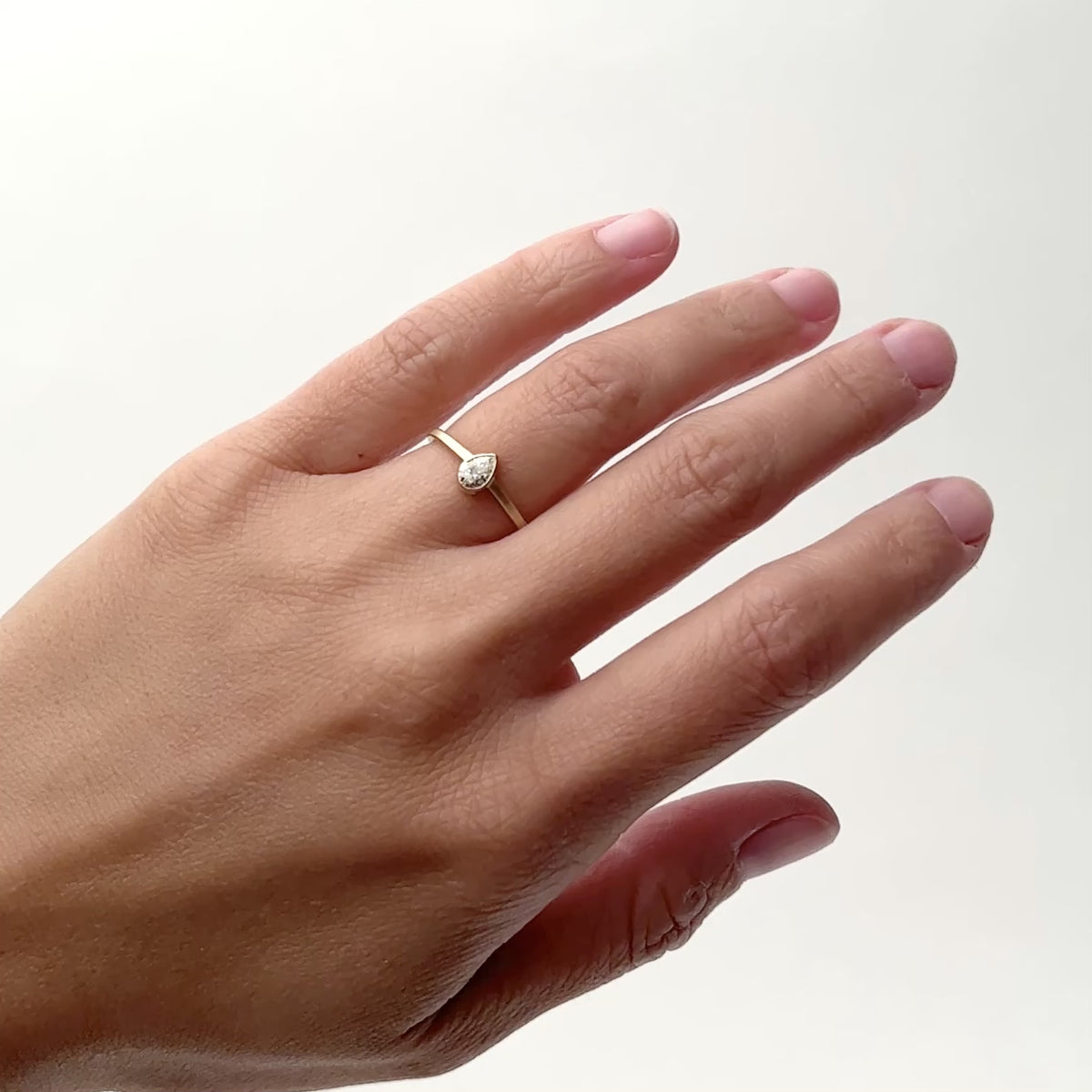 A model wears a narrow 14k yellow gold band with a bezel set .25ct pear shaped lab grown white diamond set in the center. The round portion of the setting features a cut-out. Designed and handcrafted in Portland, Oregon.