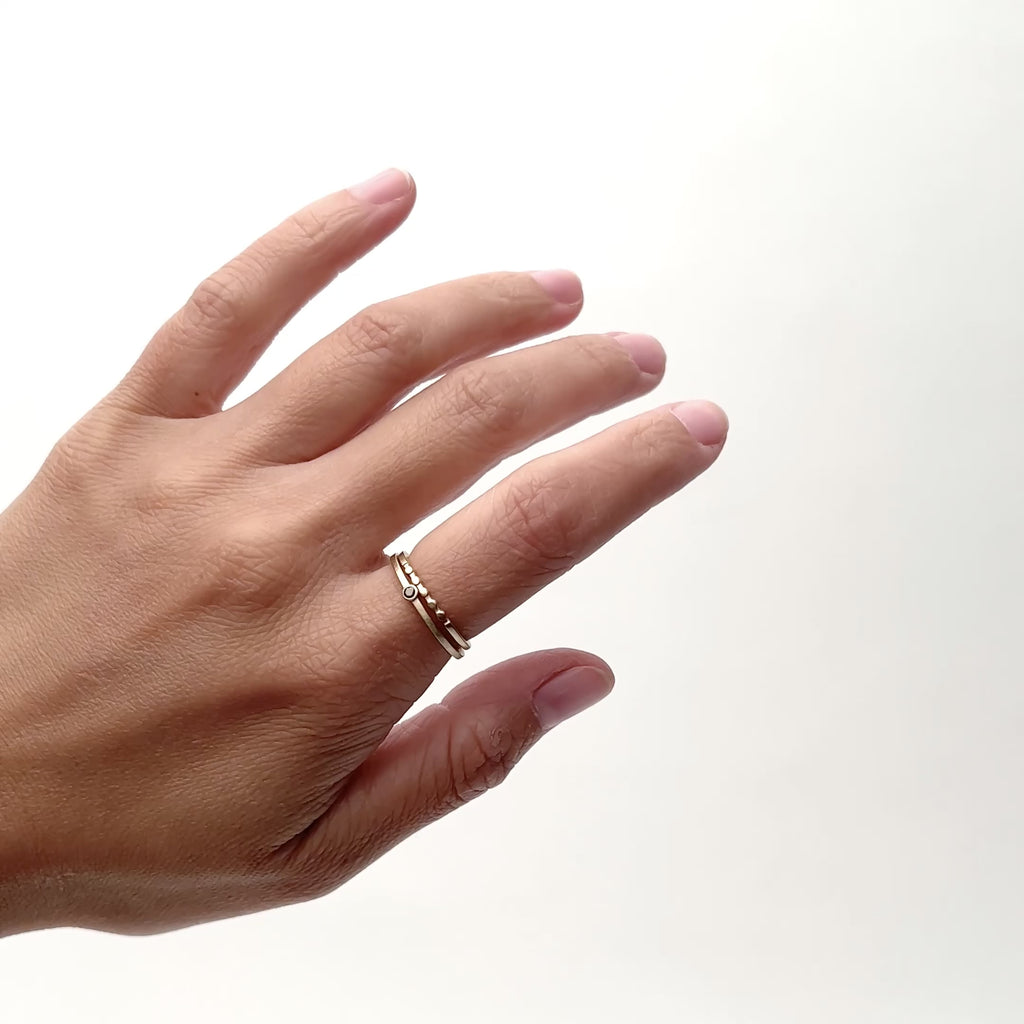 A model wears a narrow 14k gold band on their pointer finger. The ring features five circular shapes along the face of the band. Next to it is a narrow solid gold band with a petite black diamond. Designed and handcrafted in Portland, Oregon.