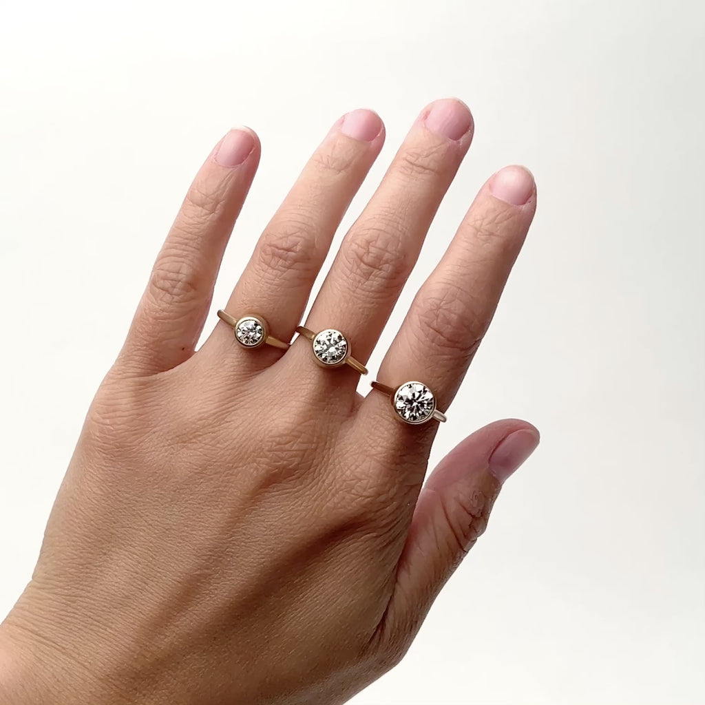 A model wears three gold rings each featuring a round white diamond. The diamond carat size varies with each ring. From left to right: 0.7 ct., 1.3 ct., and 2 ct. Each diamond is bezel set with a solid wall of gold surrounding the diamond. The band is narrow and rounded. Designed and handcrafted in Portland, Oregon.