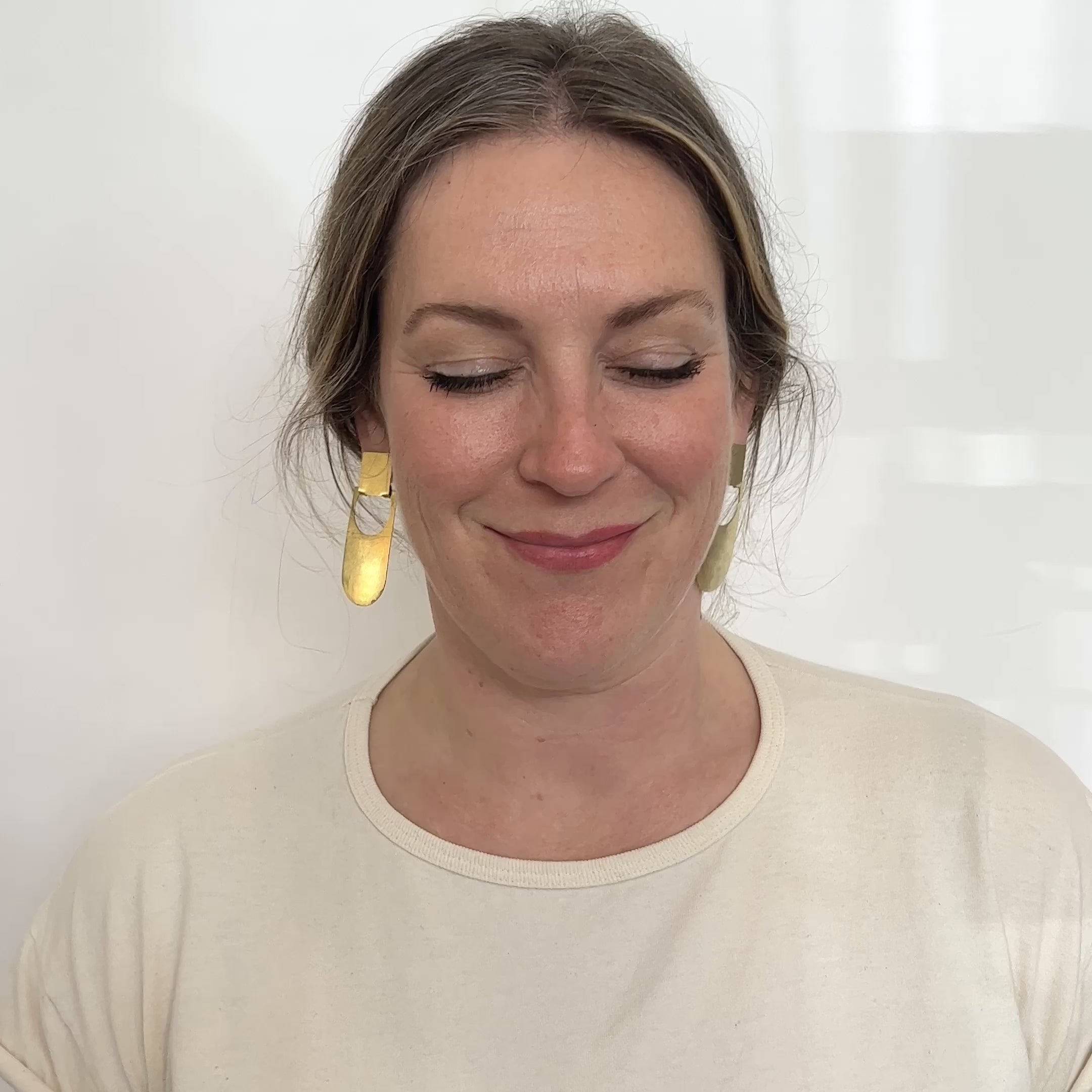 A model wears an elongated curved earring with a circular cut out that hangs from a square stud earring. They move their head side to side to show the swing of the earring. Made in brass and finished with a hammered texture. The Botao earrings are designed in Portland, Oregon.