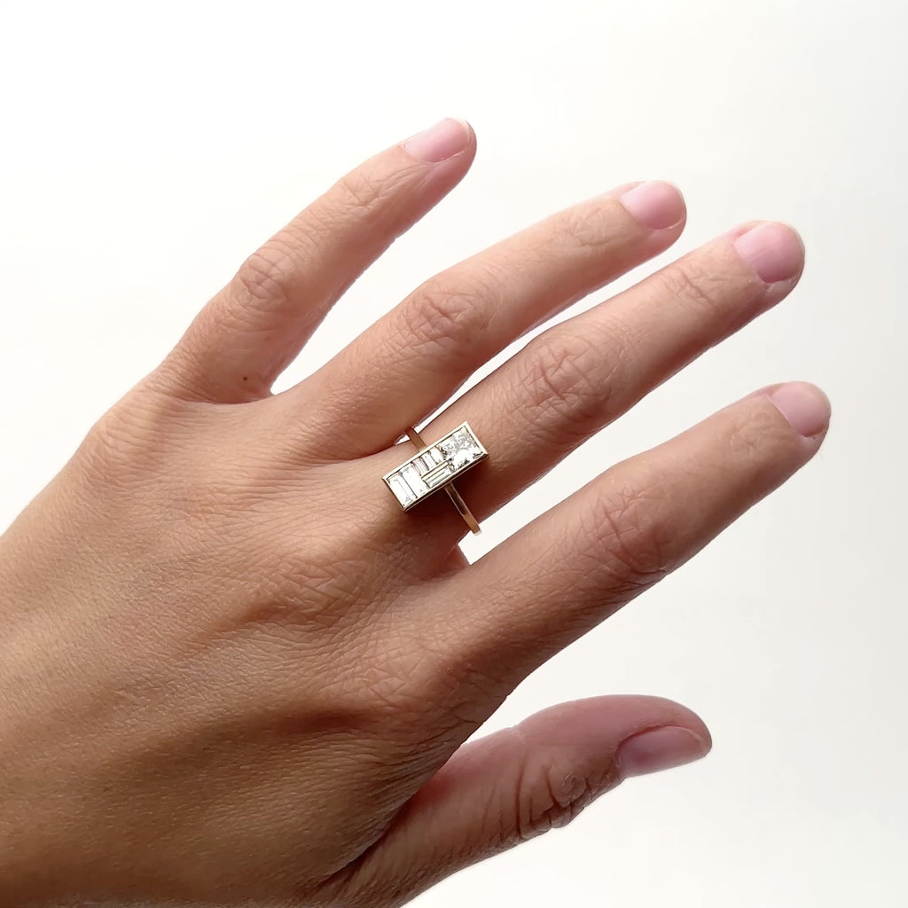 A 14k narrow band ring that features a rectangle focal piece in a north/south position. Set in the rectangle is an array of lab grown baguette diamonds and one square cut lab grown white diamond. The model wears the Mira ring on their middle finger and rotates their hand from side to side. The Miro ring is designed and handcrafted in Portland, Oregon.