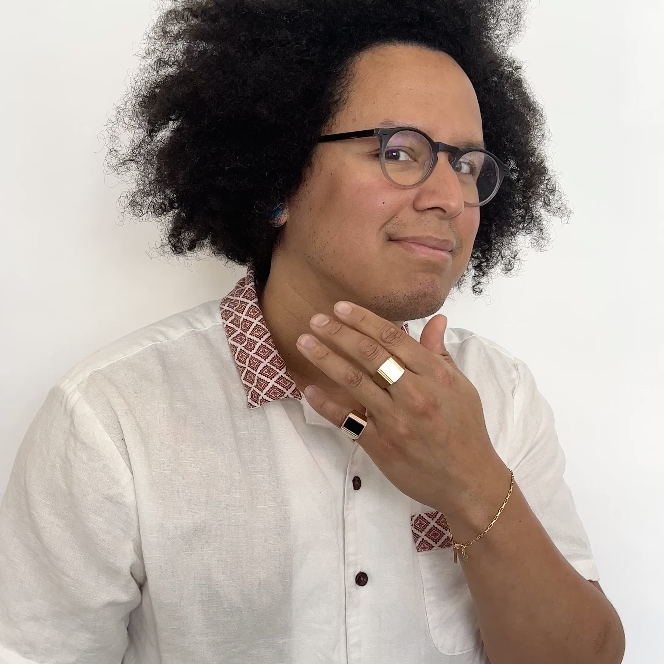 A model wears on their middle finger: a wide band rectangular signet ring with a stair step cut out design in bronze. On their pinky finger: A wide band rectangular signet ring with a stair step cut out design in bronze featuring a rectangle of Oregon black jasper. They hold their hand up to their face and wave it from side to side. Designed and handcrafted in Portland, Oregon.