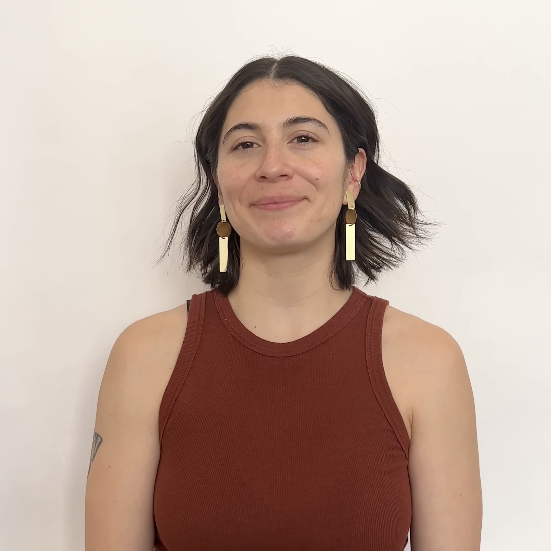 A model wears a long brass earring made with two rectangular pieces interconnected with a single circular shape. A model hakes their head from side to side to show the movement. Designed and handcrafted in Portland, Oregon.