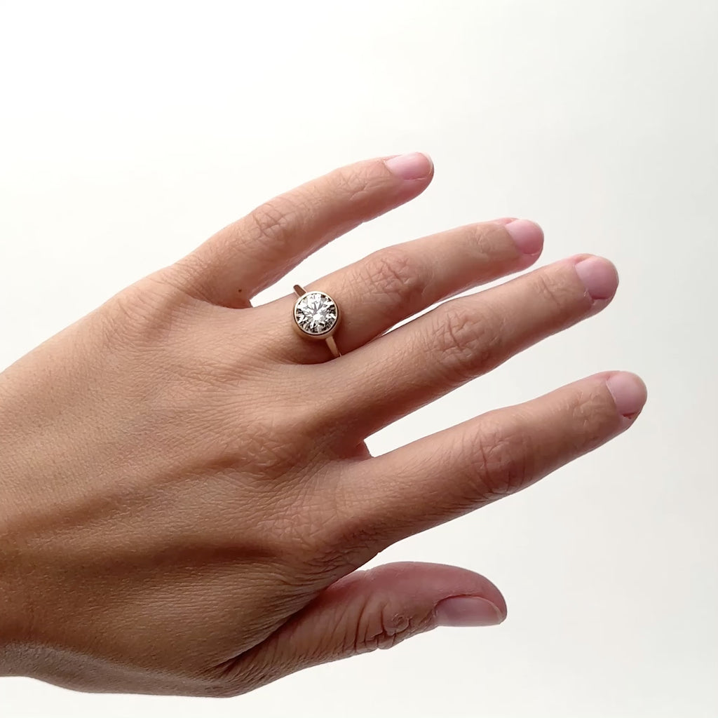 A model wears a 2 ct. diamond ring on their ring finger. The diamond is round and bezel set in 14k gold. The band is rounded and a wall of gold surrounds the sides of the diamond. Designed and handcrafted in Portland, Oregon.