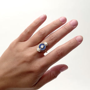 A 14k narrow band gold ring that features a central round cut lab grown white diamond with two semicircles of Chilean blue lapis. A small portion on the north and south of the ring display  five lab grown baguette diamonds with a gold cage surrounding the central focal piece. The model wears the Ara ring on their middle finger. Designed and handcrafted in Portland, Oregon.