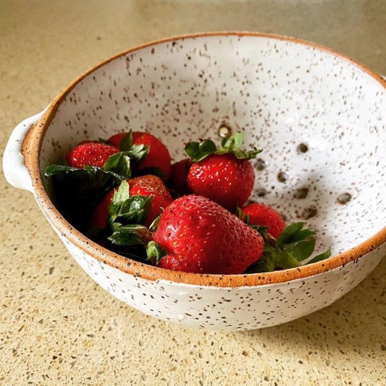 Speckled ceramic colander holds a handful of red strawberries. Glazed in a matte white finish with an exposed natural clay edge, the White Colander is designed and handmade by Byun Ceramics in Portland, OR.