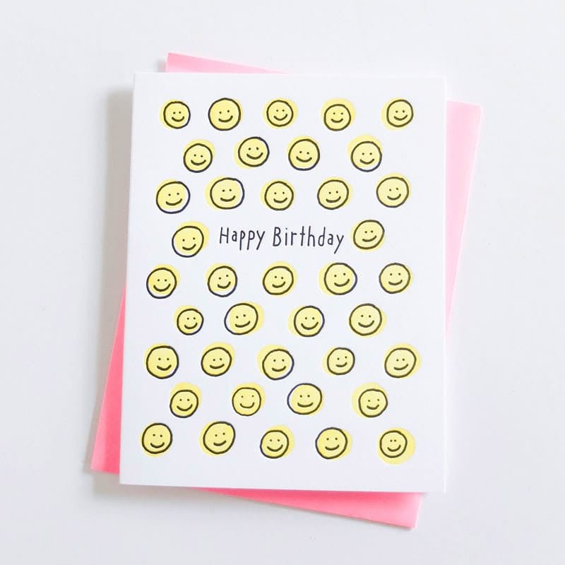 White greeting card adorned with yellow smiley faces. Center of card reads: "HAPPY BIRTHDAY" in black text. Designed by Ashkahn and printed in Portland, OR.
