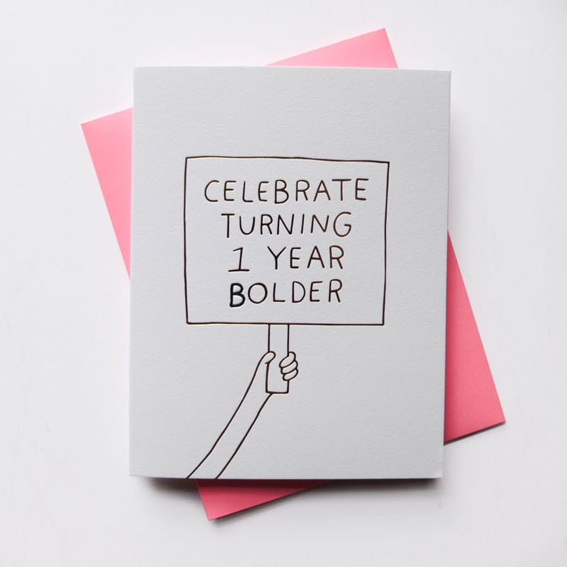 White card with an image of an arm holding up a sign that reads: "ONE YEAR BOLDER" in gold colored font. Designed by Ashkahn and printed in Portland, Oregon.