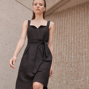 A model wears a black mid-length dress with straps and a black sash. The Aya Dress in Noir is designed by Eve Gravel and made in Montreal, Canada.