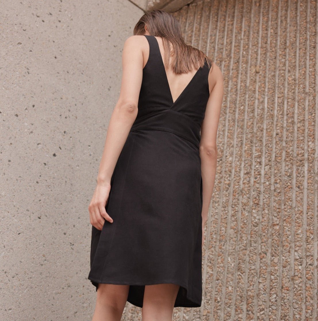A model shows the back side of a black mid-length dress with straps and a black sash. The Aya Dress in Noir is designed by Eve Gravel and made in Montreal, Canada.
