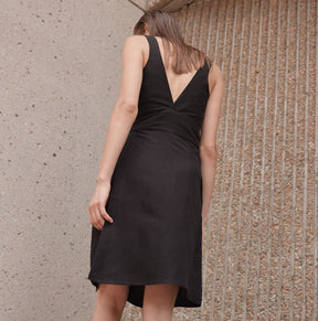 A model shows the back side of a black mid-length dress with straps and a black sash. The Aya Dress in Noir is designed by Eve Gravel and made in Montreal, Canada.