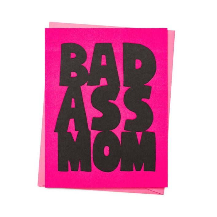 Front of card reads: "BAD ASS MOM" in black letters against a bright pink background. Comes with a pink envelope. Designed by Ashkahn and printed in Portland, OR.