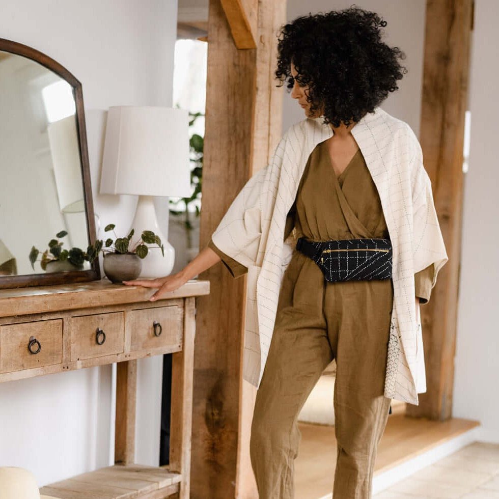 A model wears a black crossbody bag with white stitched detail around the waist. The Crossbody Belt Bag in Charcoal is designed by Anchal in Louisville, KY and handmade in Ajmer, India.