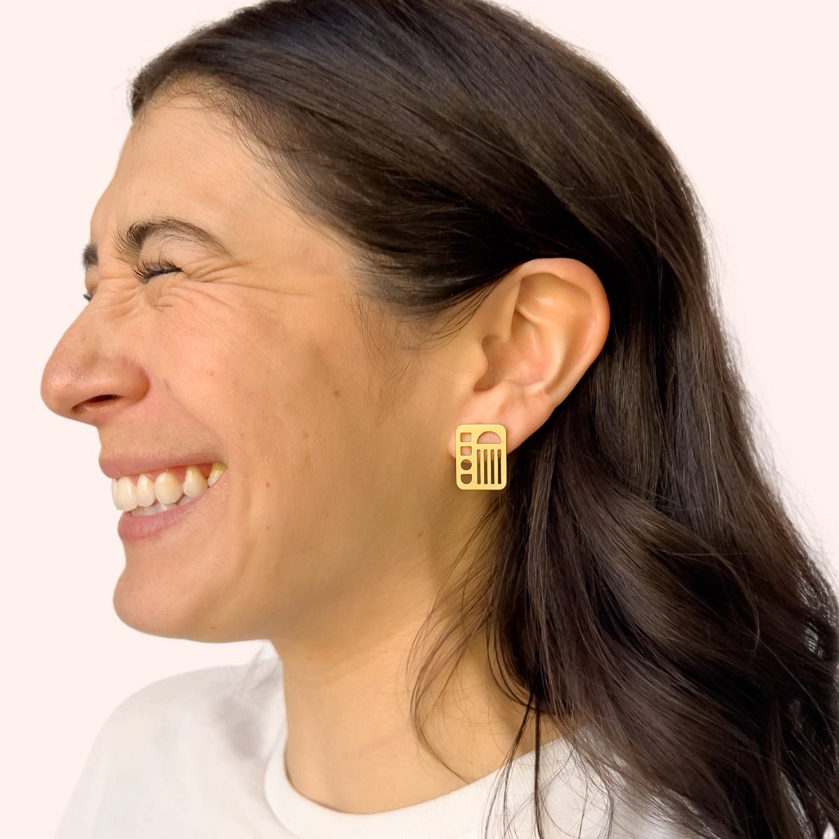 A model wears small brass rectangular stud earring with circles, lines, squares and half moon cut outs. Designed and handcrafted in Portland, Oregon.