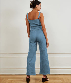 A model shows the back side of a light blue denim jumpsuit with narrow straps, flared bottom and zipper up the front. The Billy Jumpsuit in Light Indigo is designed by Loup and made in New York City, USA.