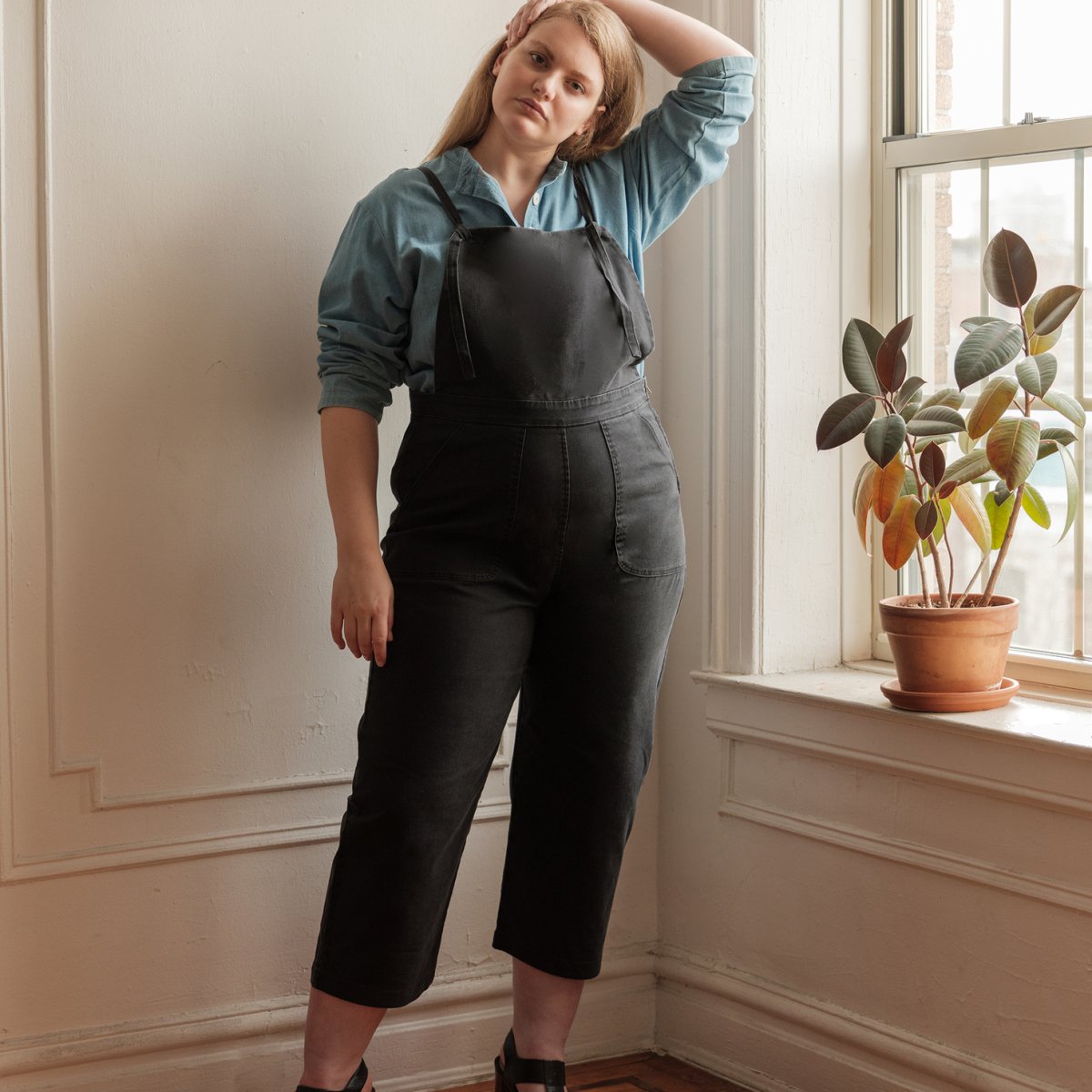Model wears cropped black overalls with thin adjustable straps and two front pockets over a blue button up shirt. The Knot Overalls in Black are designed by Loup and Made in New York City, NY.