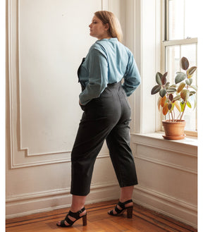 Model shows the back side of cropped black overalls. Overalls are backless with thin adjustable straps and two back pockets over a blue button up shirt. The Knot Overalls in Black are designed by Loup and Made in New York City, NY.