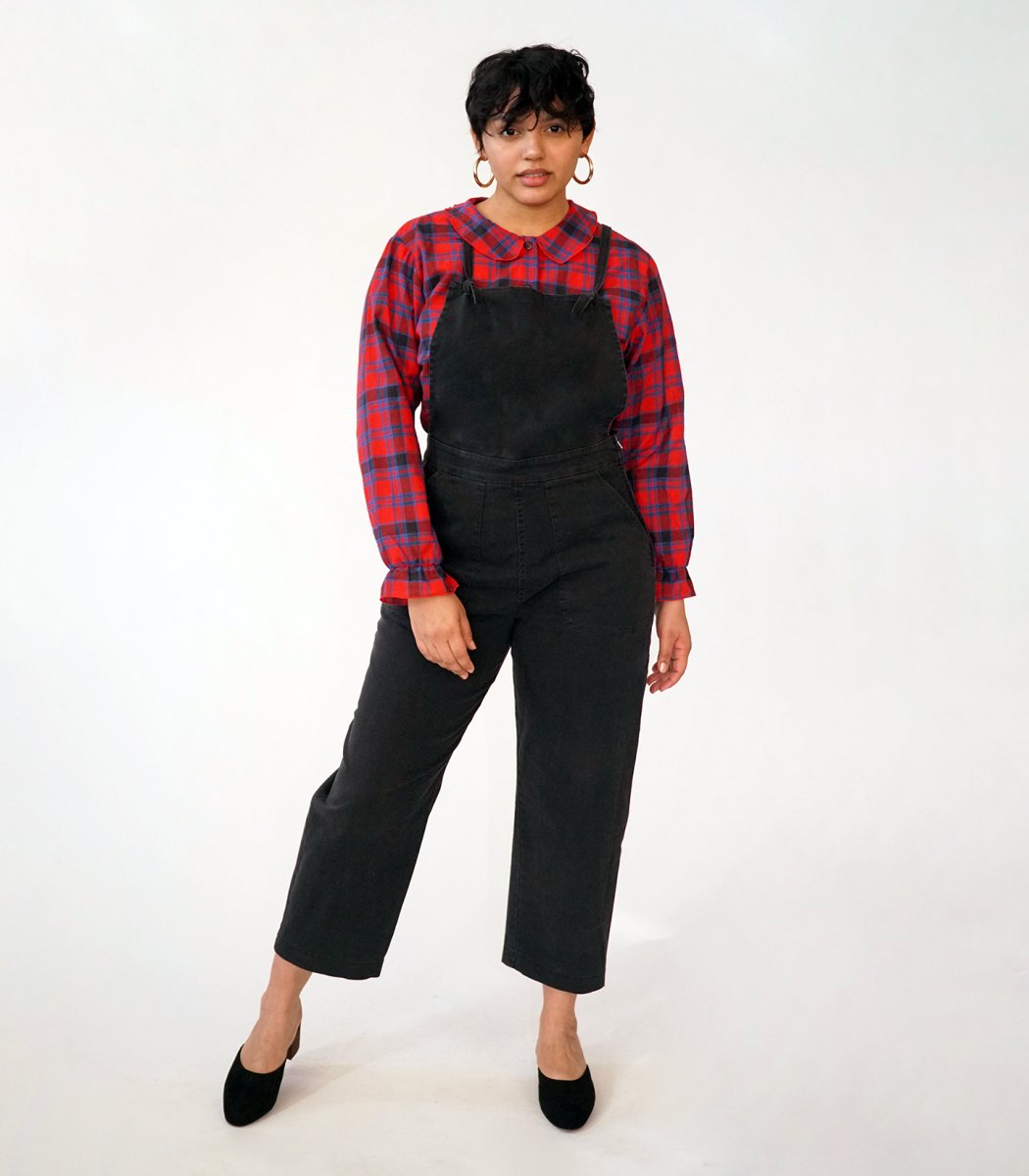 Model wears cropped black overalls with thin adjustable straps and two front pockets over a bright red plaid button up shirt. The Knot Overalls in Black are designed by Loup and Made in New York City, NY.