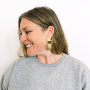 A model wears an elongated curved earring with a circular cut out that hangs from a square stud earring. Made in brass and finished with a hammered texture. The Botao Earrings are designed and handcrafted in Portland, Oregon.