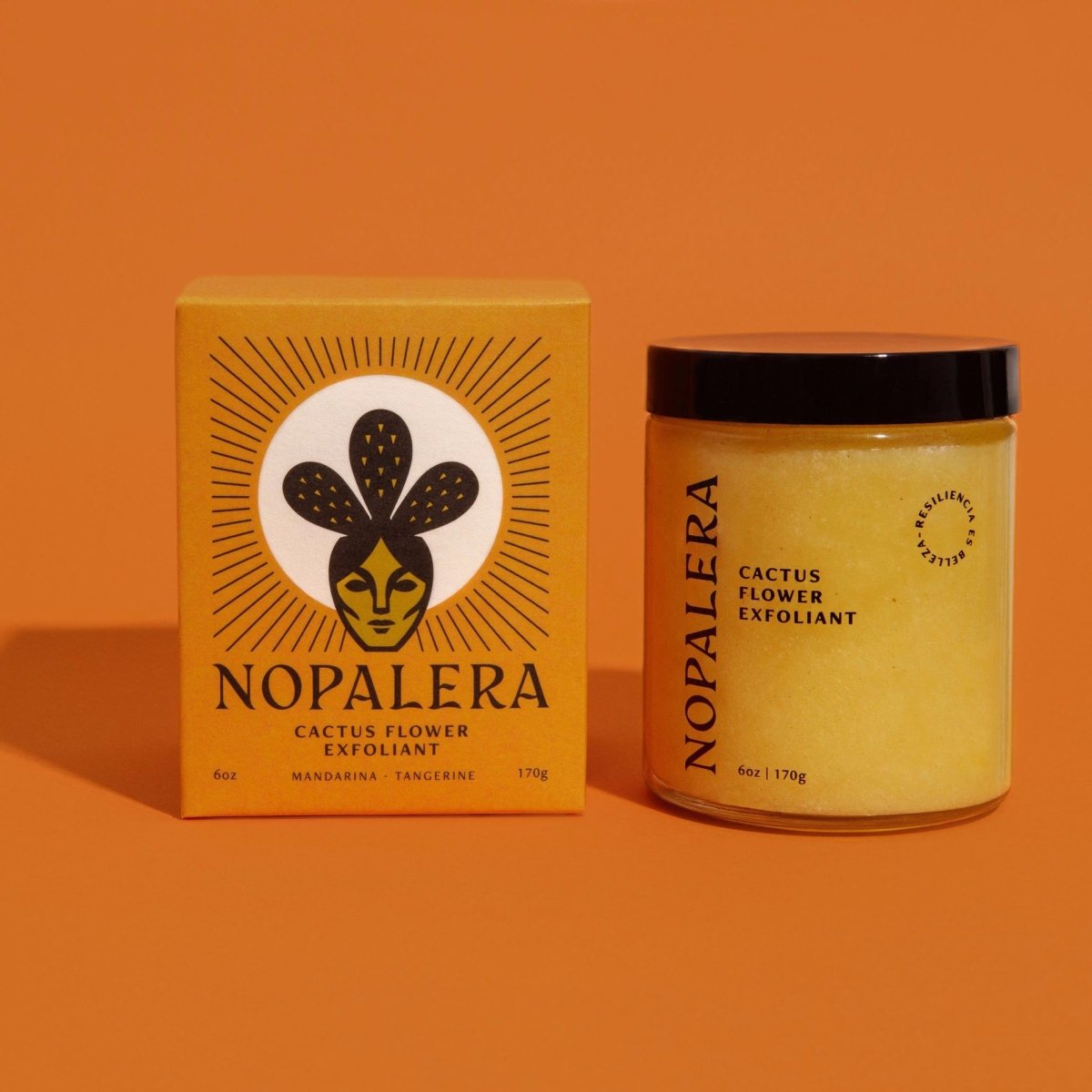 A glass jar of a yellow exfoliant mixture sits to the right of an orange box with a  cactus and facial illustration. The Cactus Flower Exfoliant is from Nopalera and made in Brooklyn, NY.