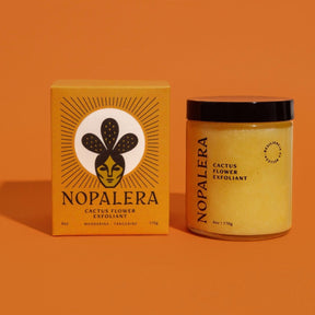 A glass jar of a yellow exfoliant mixture sits to the right of an orange box with a  cactus and facial illustration. The Cactus Flower Exfoliant is from Nopalera and made in Brooklyn, NY.