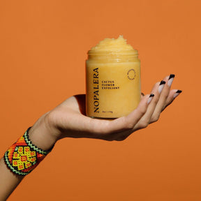 A model's hand holds out an open glass jar filled with a pale yellow exfoliating mixture. The Cactus Flower Exfoliant is from Nopalera and made in Brooklyn, NY.
