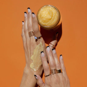 A model's hands apply a pale orange exfoliating mixture to the skin. Next to the hands is an open glass jar containing a pale yellow exfoliating mixture. The Cactus Flower Exfoliant is from Nopalera and made in Brooklyn, NY.