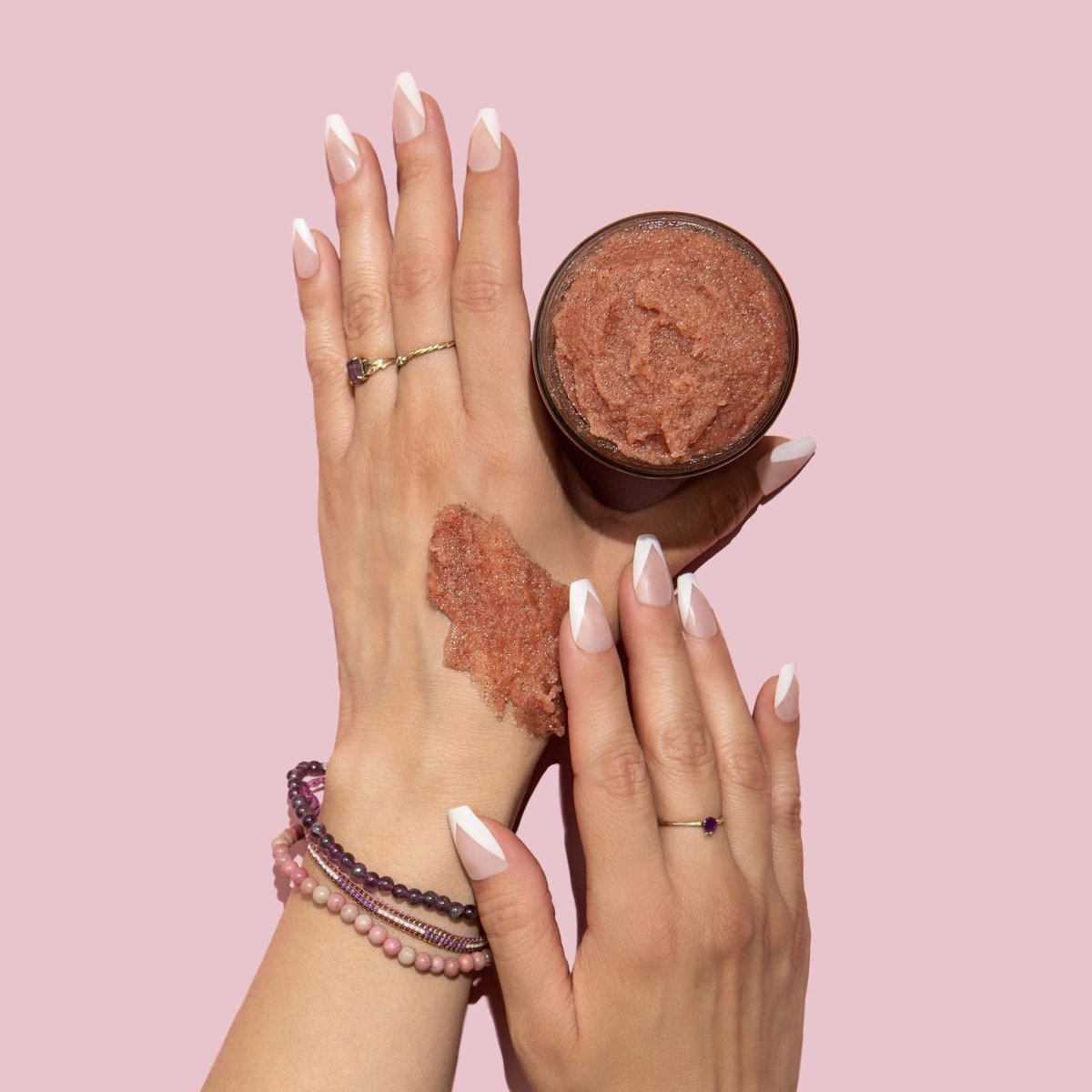A model's hands apply a pale pink exfoliating mixture to the skin. Next to the hands is an open glass jar containing a pale pink exfoliating mixture. The Cactus Flower Exfoliant is from Nopalera and made in Brooklyn, NY.
