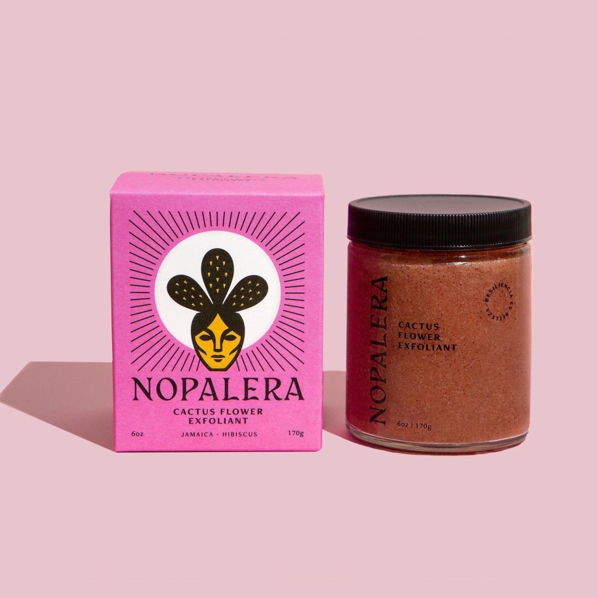 A glass jar of a pink exfoliant mixture sits to the right of a pink box with a cactus and facial illustration. The Cactus Flower Exfoliant is from Nopalera and made in Brooklyn, NY.