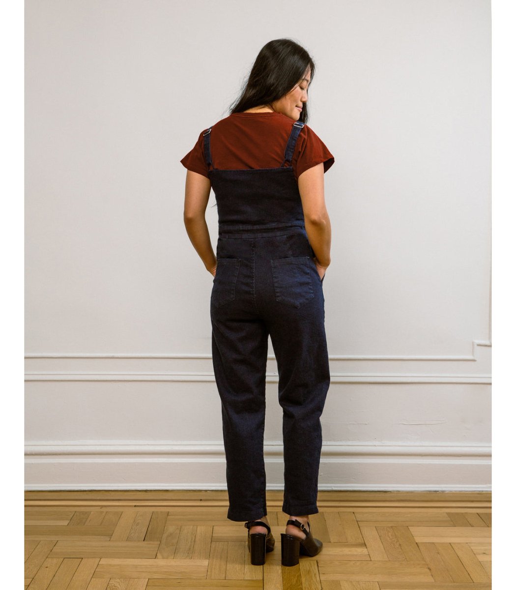 A model shows the backside of dark denim jumpsuit with adjustable straps and a front zipper. The Claudia Jumpsuit in Dark Indigo is designed by Loup and made in New York City, USA.