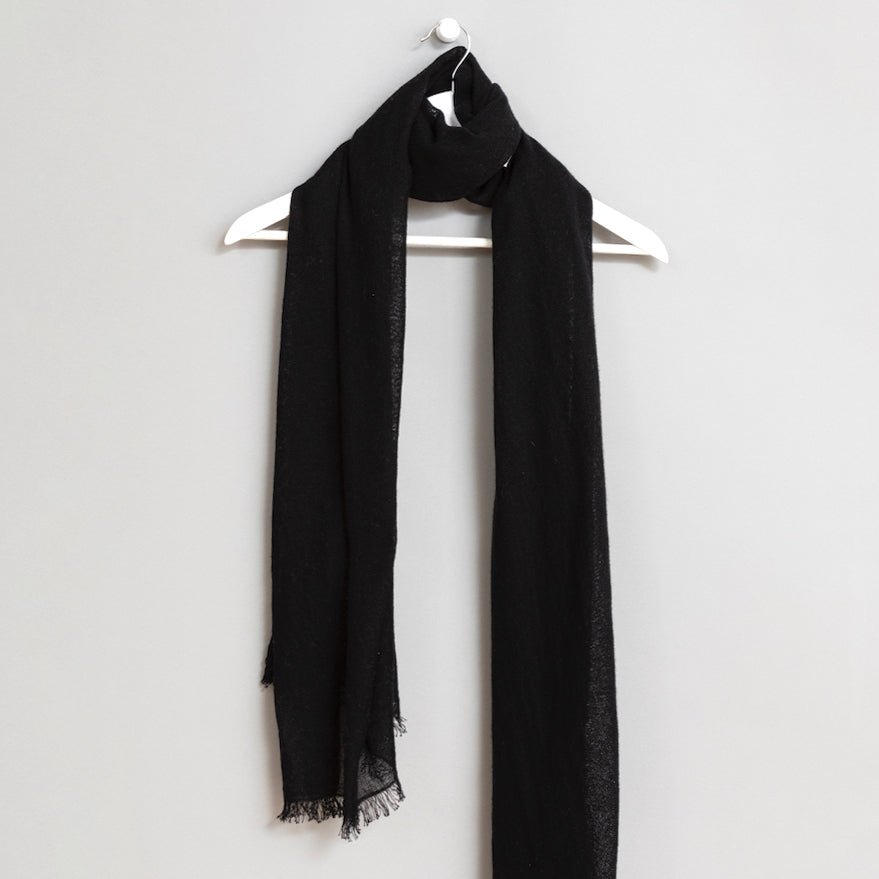 Black scarf wrapped around a white wooden hanger. The scarf has frayed edges. The Merino Woven Scarf in Black is from designer Dinadi.