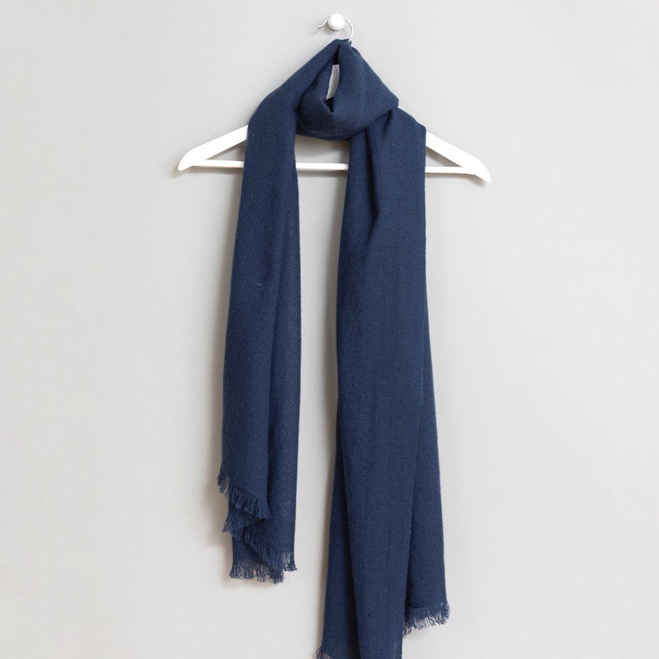 Blue scarf wrapped around a white wooden hanger. The scarf has frayed edges. The Merino Woven Scarf in Dark Blue is from designer Dinadi.