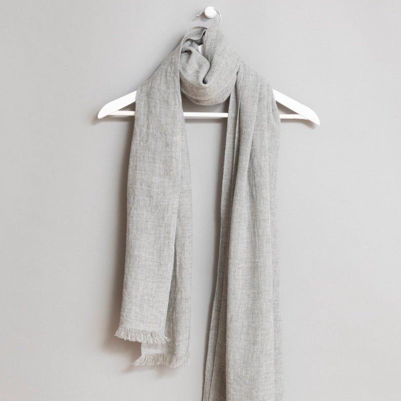 Grey scarf wrapped around a white wooden hanger. The scarf has frayed edges. The Merino Woven Scarf in Flint Grey is from designer Dinadi.