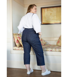 Model shows the back side of dark blue cropped overalls with thin adjustable straps over a long sleeve white shirt. Overalls are backless with two back pockets.The Knot Overalls in Dark Indigo are designed by Loup and made in New York City, NY.