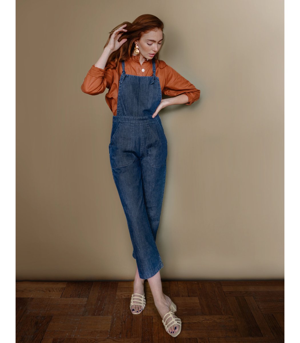 Model wears dark blue cropped overalls with thin adjustable straps and two front pockets over a long sleeve orange button up shirt. The Knot Overalls in Dark Indigo are designed by Loup and made in New York City, NY.