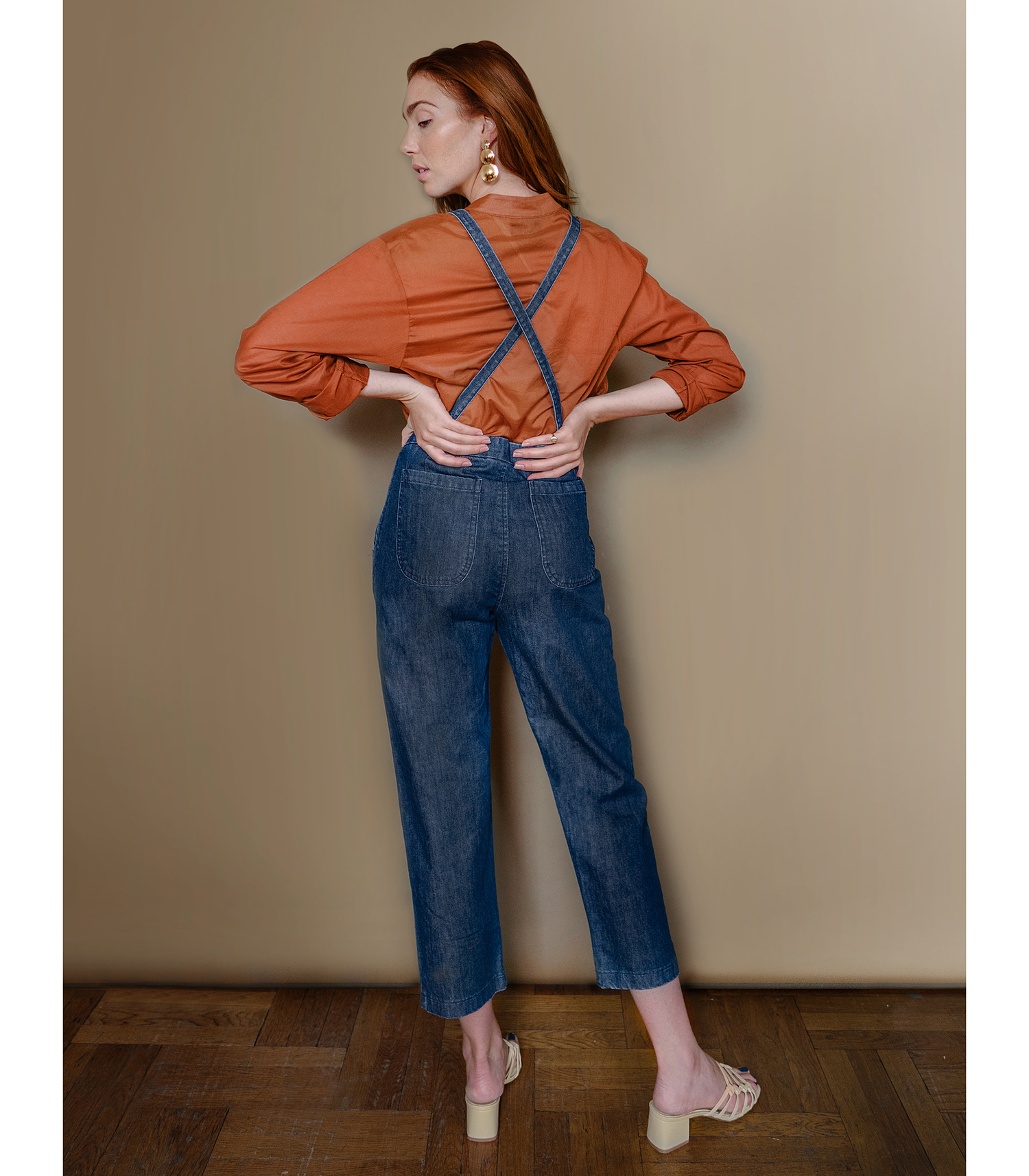 Model shows the back side of dark blue cropped overalls with thin adjustable straps and two front pockets over a long sleeve orange shirt. The Knot Overalls in Dark Indigo are designed by Loup and made in New York City, NY.
