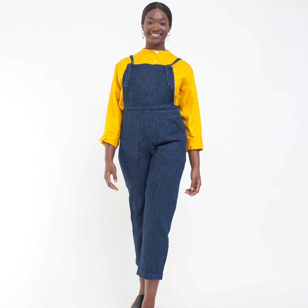 Model wears dark blue cropped overalls with thin adjustable straps and two front pockets over a long sleeve yellow shirt. The Knot Overalls in Dark Indigo are designed by Loup and made in New York City, NY.