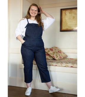 Model wears dark blue cropped overalls with thin adjustable straps and two front pockets over a long sleeve white shirt. The Knot Overalls in Dark Indigo are designed by Loup and made in New York City, NY.