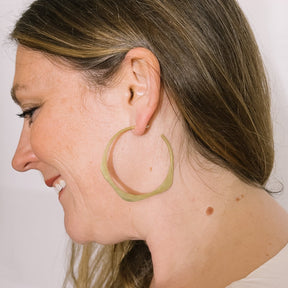 A model wears a large brass hoop earring in a wave design with a hand hammered texture and sterling silver earring posts. The large Doble Hoop Earrings are designed and handcrafted in Portland, Oregon.