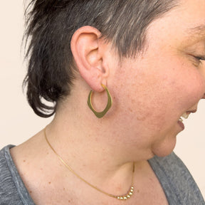 A model wears a brass hoop earring in a wave design with a hand hammered texture and sterling silver earring posts. The small Doble Hoop Earrings are designed and handcrafted in Portland, Oregon.