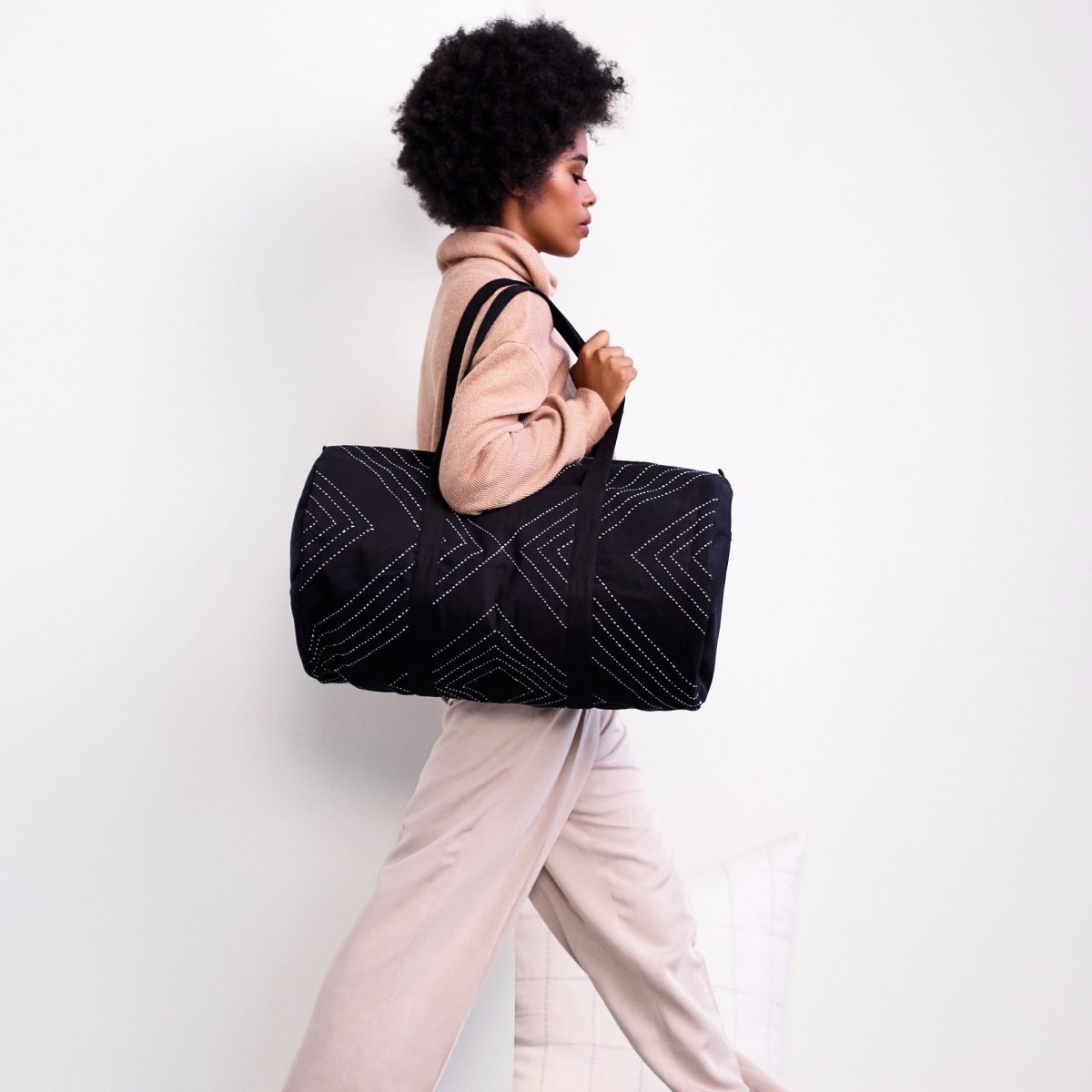 Model carries black duffel bag with white stitched geometric design. Designed by Anchal in Louisville, Kentucky and made in Ajmer, India.