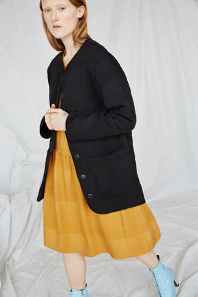 A standing red-haired woman wears a black coat and yellow dress. The Cost Jacket in Black Plaid is from Canadian designer Eve Gravel.