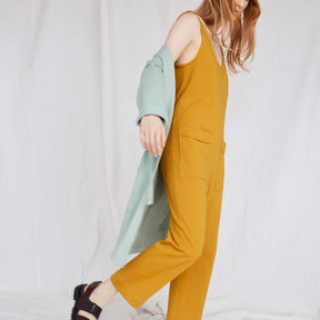Red-haired model wears yellow jumpsuit and light blue duster while moving to the right. The Owen Jumpsuit in Marigold is from Canadian designer Eve Gravel.