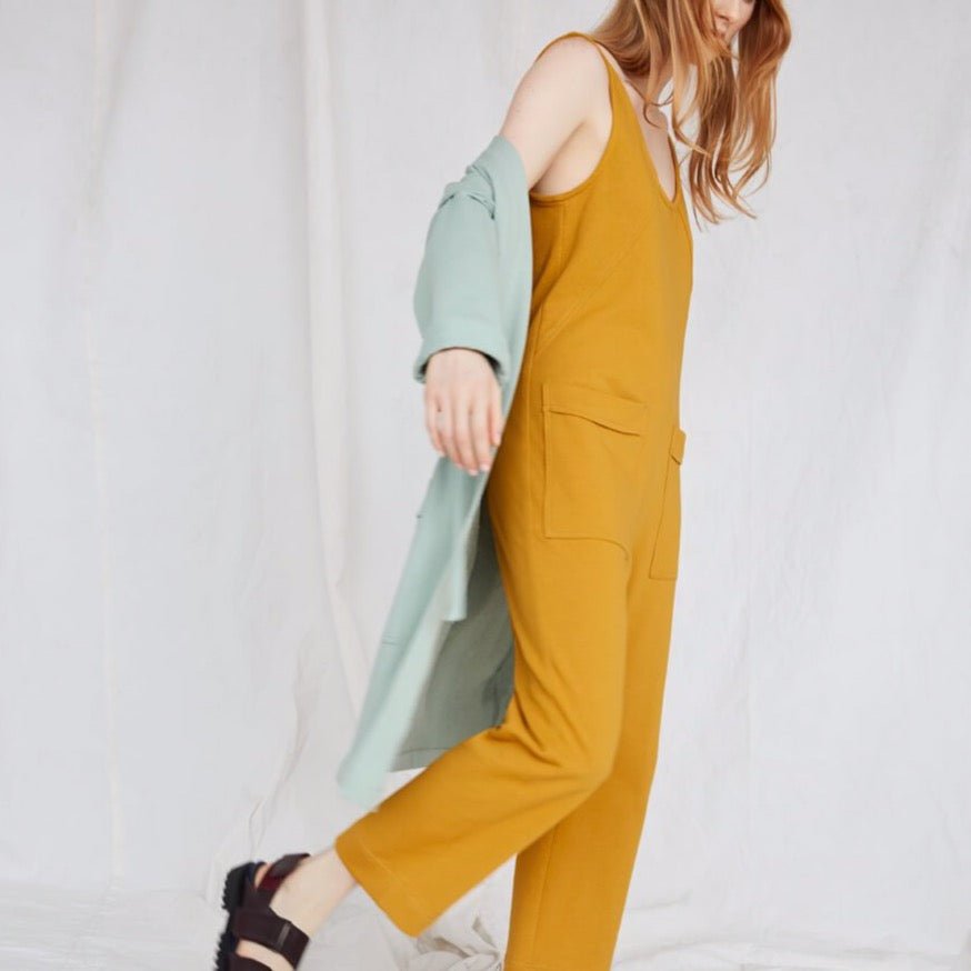 Red-haired model wears yellow jumpsuit and light blue duster while moving to the right. The Owen Jumpsuit in Marigold is from Canadian designer Eve Gravel.