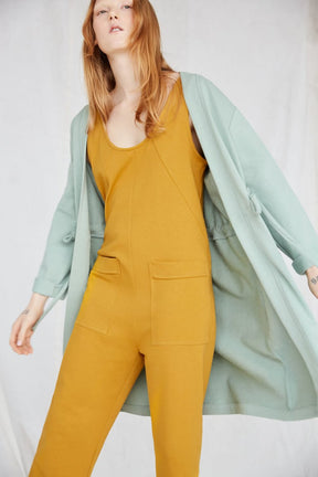 Red-haired model wears yellow jumpsuit and light blue duster while swinging her arms. The Owen Jumpsuit in Marigold is from Canadian designer Eve Gravel.