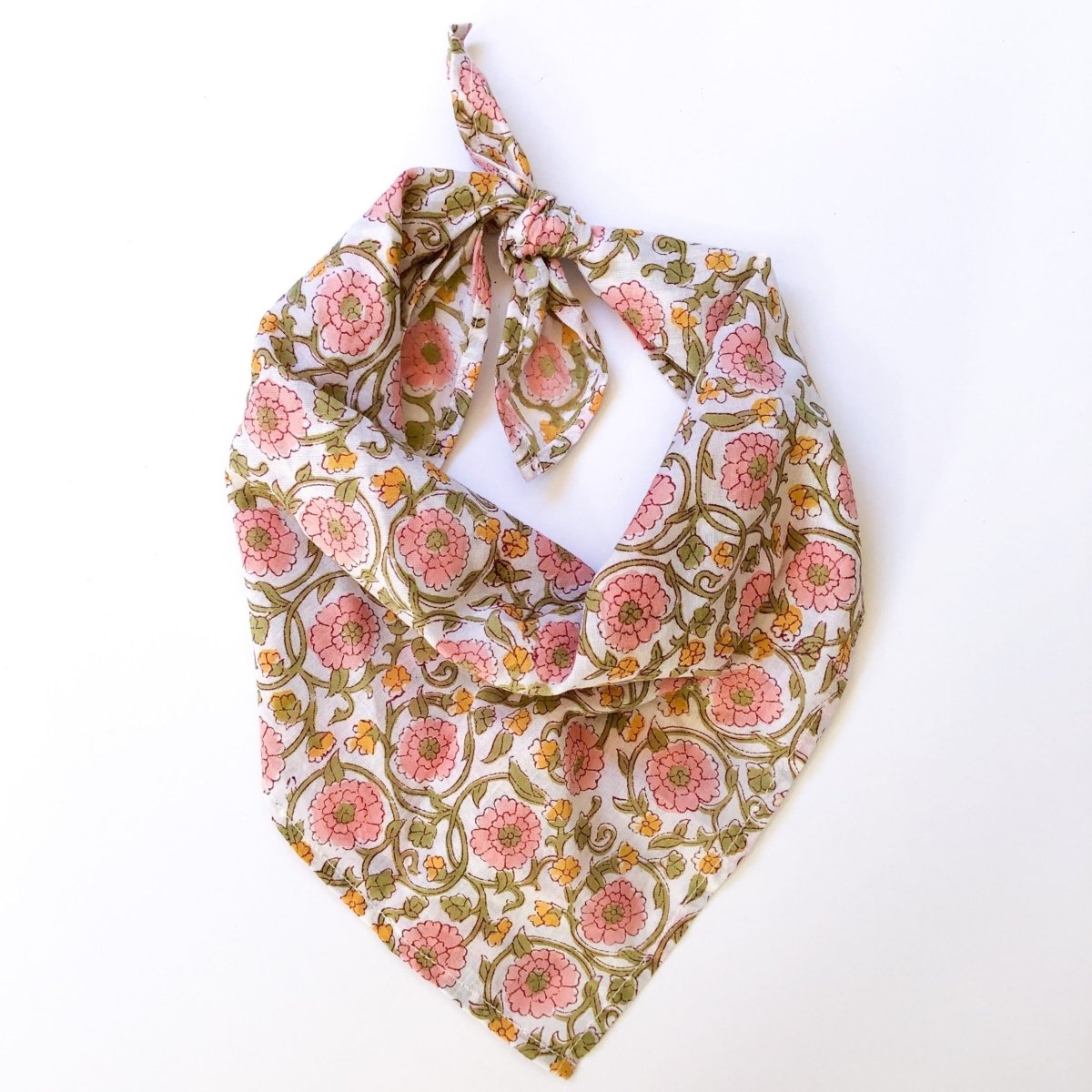 A pink, yellow, white and green floral patterned bandana, folded over and tied in a knot. Block printed by hand, the Flora Bandana from Maelu is designed in Portland, Oregon and handmade in India.