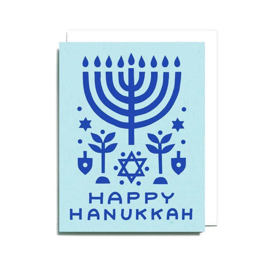 A light blue card with a dark blue festive menorah in a symmetrical paper cut collage. Front of card reads: "HAPPY HANUKKAH." Designed and handcrafted by Worthwhile Paper in Ypsilanti, MI. 