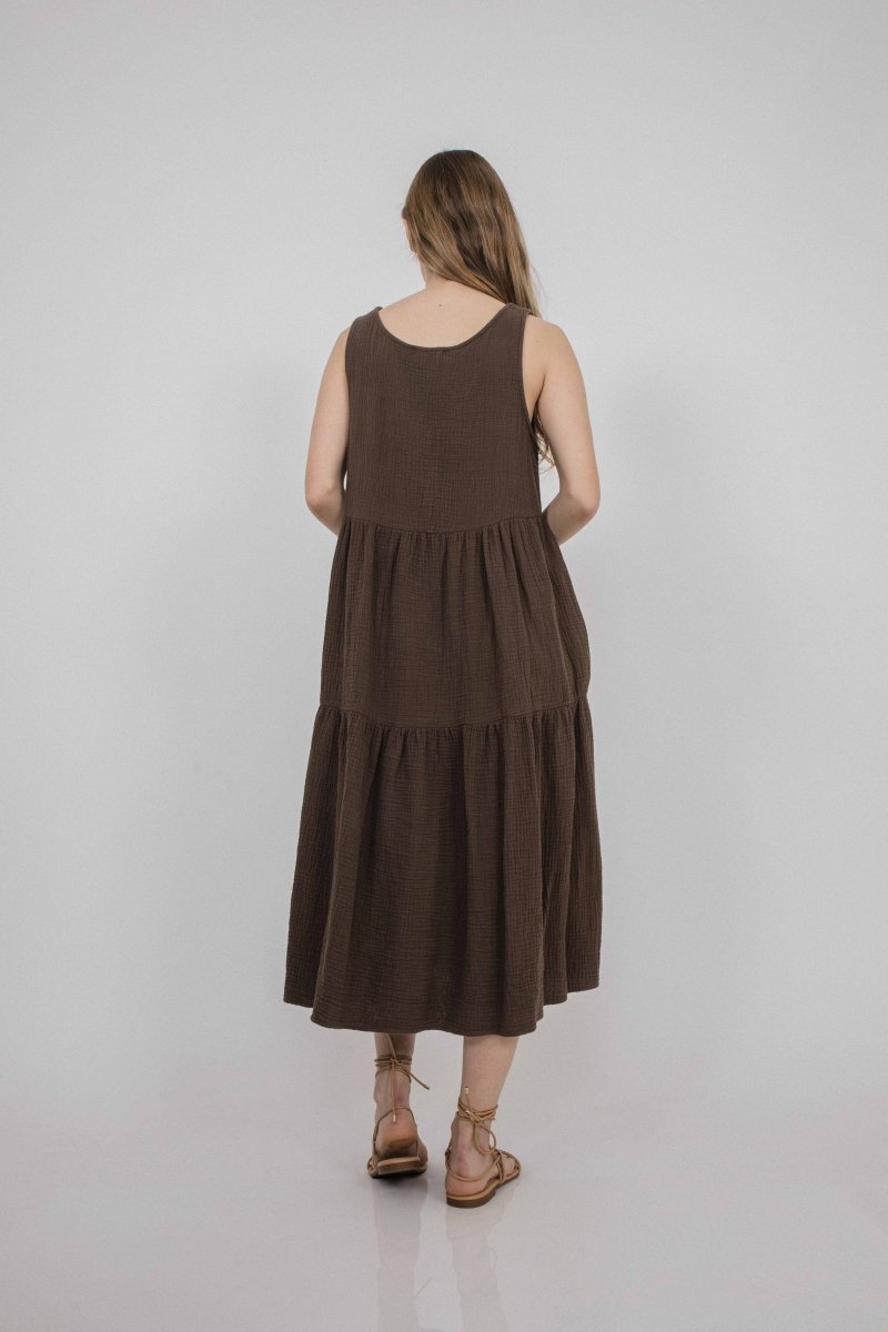 A model shows the backside of a sleeveless loose fitting dress with two tiers of fabric that form the bottom portion of the dress. The Harlow Dress on Cocoa is designed and made by Corinne Collection in Los Angeles, CA