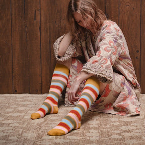 A model wears a knee high striped sock with yellow, red, pink and blue stripes. The toe and heel are a mustard yellow color. The Hazel Knee High Sock in Goldenrod Multi is from Zkano and made in Alabama, USA.