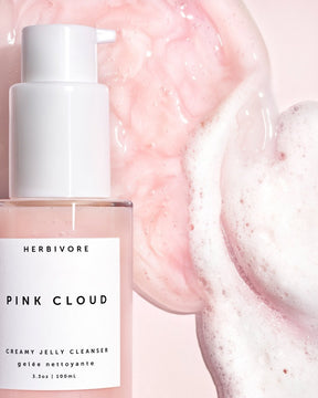 Closeup of a glass bottle with a pink foaming cleanser poured next to it.  The Pink Cloud Cleanser is from the brand Herbivore.